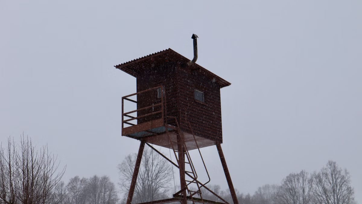A wooden watchtower stands tall amidst falling snow and barren trees, creating a serene winter forest scene.